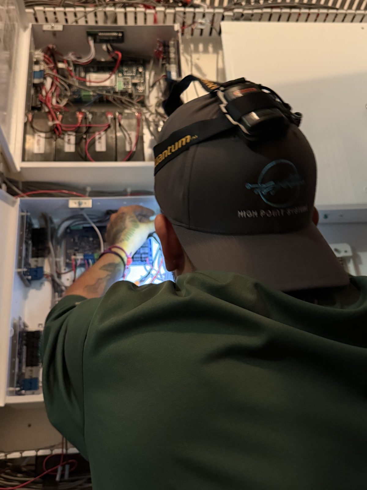 HPSI technician in a branded cap working inside a security panel with a headlamp